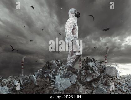 Überlebende mit weißer Gesamt- und Gasmaske erreichen die Spitze der Müllhalde und beobachten den apokalyptischen Himmel Stockfoto