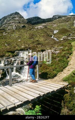 Weibliche Wanderer auf Holzfußbrücke über tumbling Bergbach mit dem Gipfel von Trolamul oberhalb, Gleann Trolamaraig, Insel Harris, Schottland, Großbritannien. Stockfoto