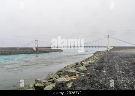 Der Kanal Joekulsarlon Glacier Lagoon überbrückte durch Hängebrücke, die Eisberge in Richtung Diamond Beach in Island spülte Stockfoto