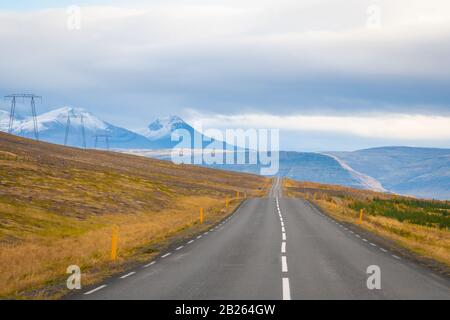 Straßenfahrt in Island leere gepflasterte Straße führt über Berge mit schneebedeckten Spitzen neben Stromleitungen Stockfoto
