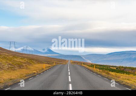 Straßenfahrt in Island leere gepflasterte Straße führt über Berge mit schneebedeckten Spitzen neben der Stromleitung im Herbst Stockfoto