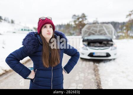 Gestresste Frau und zerbrochenes Auto mit geöffneter Haube im Hintergrund. Probleme bei der Fahrt und Konzepte der Hilfe. Stockfoto