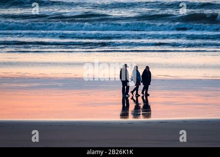 Die Silhouette von 3 Einzelreisenden, die am Strand spazieren, mit den Wellen, WT-Sandreflexionen mit Reflexionen und Farbe der Sonne auf dem Sand Stockfoto