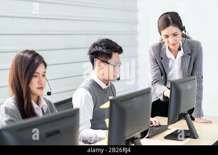 Vertrauen junger Erwachsener mit lächelnder Vorgesetzter, die vor dem Arbeitstisch des Operatorteams stehen, in ein Callcenter, um ihren Mitarbeiter zu überwachen. U Stockfoto