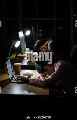Müde junge asiatische Geschäftsfrau und Geschäftsmann schlafen spät in der Nacht am Computerschreibtisch im Büro. Stockfoto