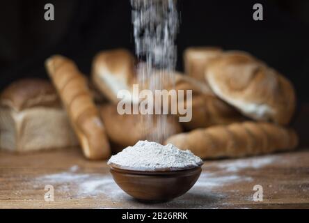 Mehl wird auf Holztisch in die Schüssel gegeben, mit verwischtem, unterschiedlichem Brot im Hintergrund. Stockfoto