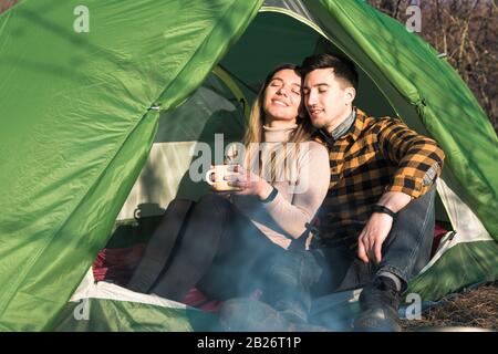 Fröhliches junges Paar, das im Freien campt, im Zelt sitzt und den Sonnenuntergang genießt Stockfoto