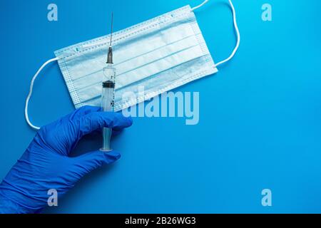 Tragen Sie einen medizinischen Handschuh ein, der eine Spritze mit einer medizinischen Impfstoffmaske auf blauem Hintergrund hält. Der Impfstoff für Coronavirus Konzept Stockfoto