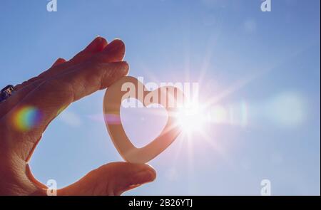 Nahaufnahme der Hand der Frau, die weißes Holzherz gegen die Sonne hält, und des blauen Himmels mit Linsenflauen und Sonnenfotografie mit Sterneneffekt. Positive Emotionen. Stockfoto