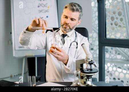 Kaukasianer Virologe, Biochemiker, Wissenschaftler, der einen weißen Mantel trägt, der das Reagenzglas hält, und die rote Flüssigkeit auf den Kolben fallen ließ. Der Chemiker untersucht den chemischen Test Stockfoto