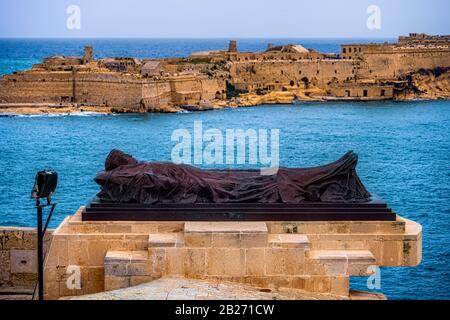 Liegeradskulptur im Belagerung Bell war Memorial in Valletta, Malta, Fort Ricasoli in Kalkara im Hintergrund Stockfoto