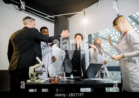 Fröhliche multiethnische Wissenschaftler Ärzte arbeiten mit Laborgeräten und stellen sich dabei gegenseitig fünf hoch. Wissenschaftlicher Erfolg Stockfoto