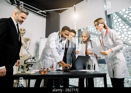 Multiethnisches Team von Ärzten, Wissenschaftlern, Mitarbeitern im Gesundheitswesen, die eine Diskussion führen, während sie Laptop verwenden, über die Ergebnisse klinischer Studien sprechen oder Stockfoto