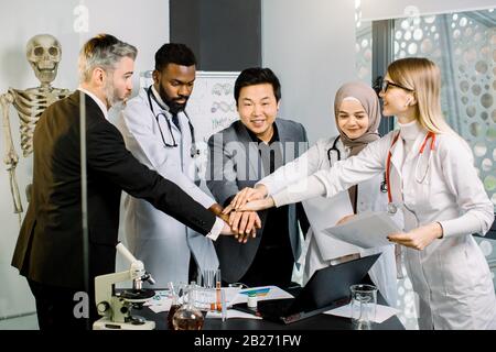 Aufnahme von multiethnischen medizinischen Teams, Ärzten, Wissenschaftlern und Laborforschern, die die Hände zusammenstapeln und sich im modernen Labor treffen. Laptop, Mikroskop und Stockfoto