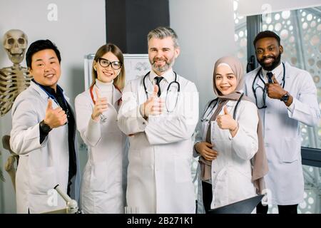 Porträt eines männlichen reifen Arztes mit multiethnischem Ärzteteam, lächelnd und mit Daumen nach oben, vor modernem Labor und menschlichem Skeletthintergrund. Wissenschaft Stockfoto