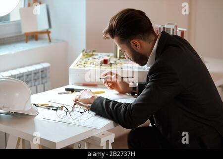 Ambitionierter aktiver Mann zeichnet Modell auf dem Papier, Nahaufnahme Foto: Interesse, Hobby, Brille, Blaupause, Stift und Schutzhelm sind auf der Th Stockfoto