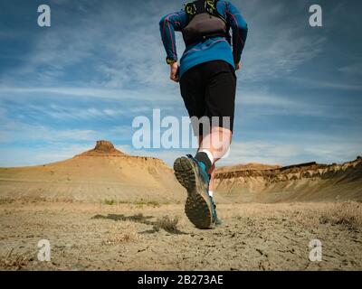 Runner Athlet mit Rucksack, der auf dem wilden Pfad in den roten Bergen der Wüste läuft. Nahansicht von hinten. Die Felsvorsprünge des Ustyurt-Plateaus Stockfoto