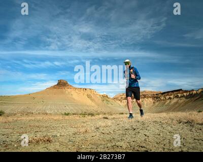 Runner Athlet mit Rucksack, der auf dem wilden Pfad in den roten Bergen der Wüste läuft. Die Felsvorsprünge des Ustyurt-Plateaus Stockfoto