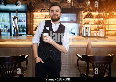 Eleganter positiver junger bärtiger Ladenassistent, der ein Glas Kakao hält. Mann genießt Trinklatte, Nahaufnahme Stockfoto
