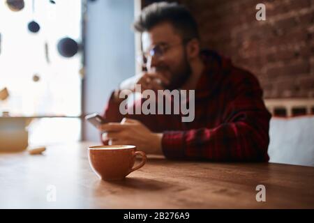 Lächelnder glücklicher Mann wird Freude daran, im verschwommenen Hintergrund des Fotos mit einem Freund zu plaudern und sich auf die Tasse Tee zu konzentrieren Stockfoto