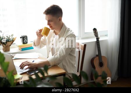 Junger, gut aussehender Mann, der am Tisch sitzt und Kaffee trinkt, Film und Film sieht. Nahaufnahme Foto. Entspannung. Kerl mit Frühstück im w Stockfoto