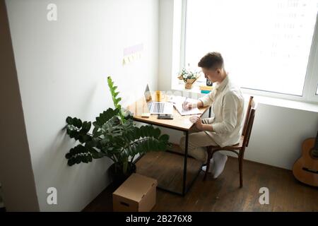 Konzentrierter, hart arbeitender junger blonder Mann mit Laptop und Notizen zu Hause im Wohnzimmer, Kerl schreiben Lebenslauf, Seitenansicht Foto. Stockfoto
