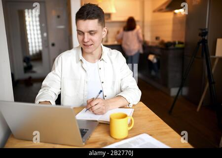 Gutaussehender blonder Geschäftsmann, der seinen Arbeitsprozess zu Hause genießt, während seine Frau das Frühstück kocht, Familie, Freizeit, Lifestyle. Stockfoto