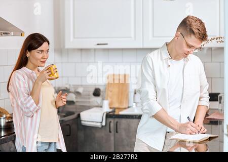 Tolle junge schöne Frau trinkt e, während ihr Freund an einem Projekt arbeitet, Nahaufnahme Foto. Beziehung, Kerl mit Stift auf seinem Tagebuch Stockfoto