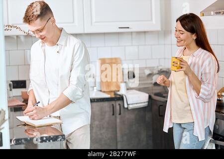 Angenehme Frau lacht über ihren Freund, der einen Plan aufstellt, schreibt seine Ideen, plant seinen Tag, Urlaub Stockfoto