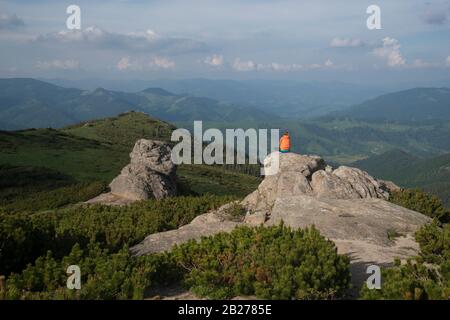 Mädchen in orangefarbener Jacke sitzt auf einem Felsen und meditiert, wobei sie die Landschaft betrachtet. Stockfoto