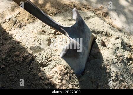 Verrosteter Spaten oder Schaufel in einer Zement- und Sand-Trockenmischung, bereit für Mörtel, mit Schatten von Ästen von einem nahe gelegenen Baum an einem sonnigen Tag Stockfoto