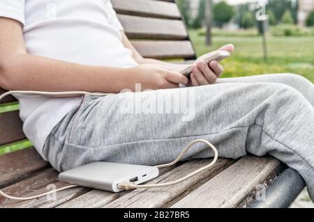 Der Junge verwendet das Telefon auf der Bank, während er von der Strombank aufgeladen wird. Stockfoto