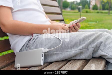 Der Junge verwendet das Telefon auf der Bank, während er von der Strombank aufgeladen wird. Stockfoto