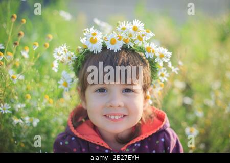 Ein kleines Mädchen im Kranz der Gänseblümchen lächelt und sitzt im Sommer im grünen Gras. Sonniger Tag. Stockfoto