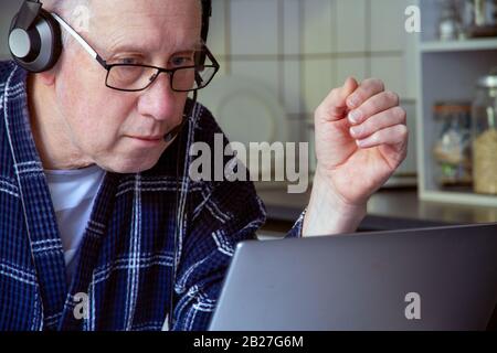 Ein älterer weißer Mann in seinem Morgenmantel und mit Kopfhörern sitzt vor einem Laptop in einer Küchenecke. Stockfoto