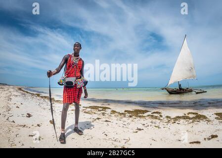 Diani Beach/Kenia - 01. März 2020: Masai am Strand in Kenia, Diani Beach, watamu hält einen Pfeil mit einem Segelboot im Hintergrund Stockfoto