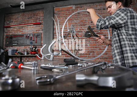Mann repariert das Fahrrad in der Werkstatt auf der Werkbank mit Werkzeugen, mit der Bürste am Rad, heimwerkerkonzept, rotem Backstein Hintergrund Stockfoto