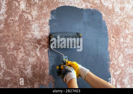 Lackierung der Wand mit einem walzengrauen Lack, Lackierung mit gelben Arbeitshandschuhen, Reparatur von Lackierwerkzeugen. Stockfoto