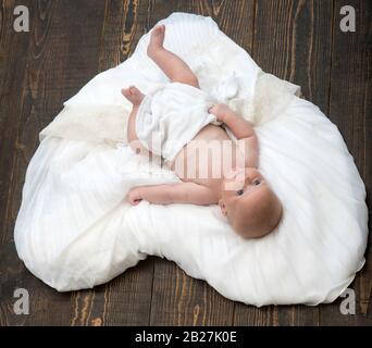 Baby liegt auf weisser weicher Federdecke, die als Herz konzipiert ist. Kleinkind mit weißer Decke bedeckt. Neugeborenes Kleinkind mit blauen Augen und ruhigem Gesicht auf Holzhintergrund. Kindheits- und Göttliches Geschenkkonzept Stockfoto