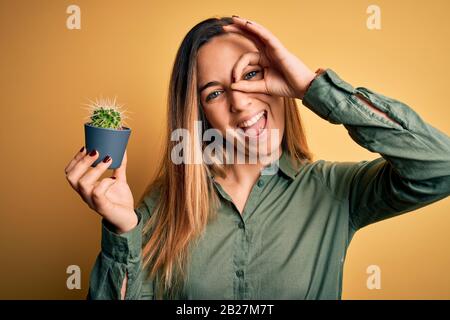 Junge, schöne blonde Frau mit blauen Augen, die einen kleinen Kakteentopf über gelbem Bckgrund hält, mit einem fröhlichen Gesicht, das lächelt, und einem guten Schild mit Hand auf dem Auge Stockfoto