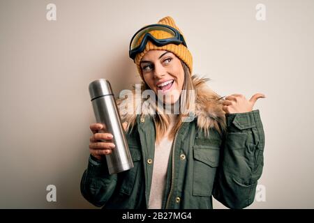 Junge schöne Brunette-Skifahrerin, die eine Skibrille trägt, die Thermo mit einem SMI trinkt, der mit dem smi mit dem Daumen zur Seite zeigt Stockfoto