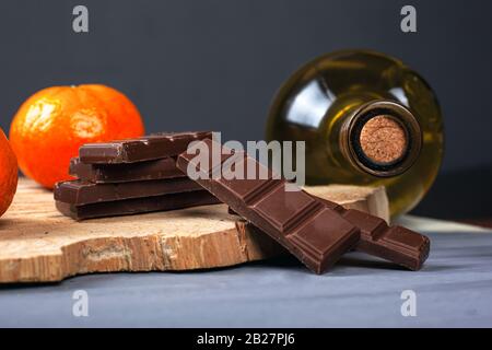 Weißweinflasche mit Milchschokolade und Mandarinen auf einem Holz-Waldregal auf grauem dunklem Hintergrund. Romantisches Abendessen für zwei Nahessen. Stockfoto