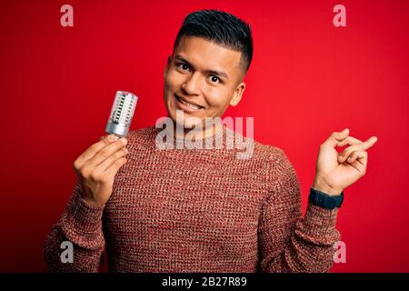 Junger gutaussehender lateinischer Mann, der die Glühbirne über dem isolierten roten Hintergrund hielt, sehr glücklich, mit der Hand und dem Finger zur Seite zu zeigen Stockfoto