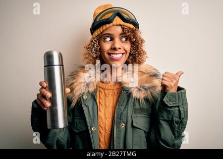 Junge afroamerikanische Skifahrerin, die eine Skibrille trägt, die Thermo mit Kaffee trinkt und mit Daumen nach oben zeigt, mit fröhlichem Gesichtsschmierl Stockfoto
