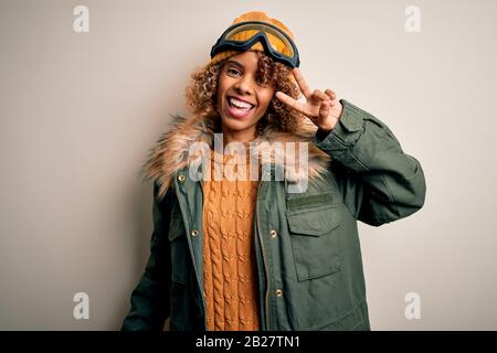 Junge afroamerikanische Skifahrerin mit lockigen Haaren, die Schneesportkleidung trägt und eine Skibrille, Die Mit den Fingern über dem Gesicht ein Symbol für Frieden macht, fröhlich lächelt Stockfoto