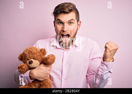 Junger blonder Mann mit Bart und blauen Augen, der Teddybärspielzeug über rosafarbenem Hintergrund hält und stolz schreit und Sieg und Erfolg sehr aufgeregt feiert, Stockfoto