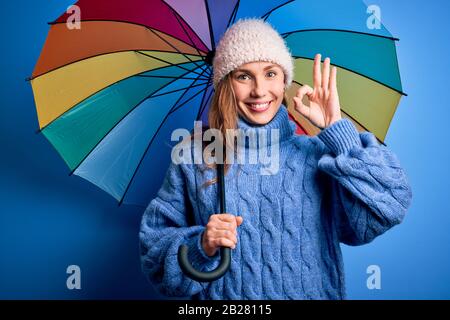 Junge, schöne blonde Frau, die einen farbenfrohen Regenschirm über einem isolierten blauen Hintergrund hält und mit den Fingern ein gutes Zeichen macht, hervorragendes Symbol Stockfoto