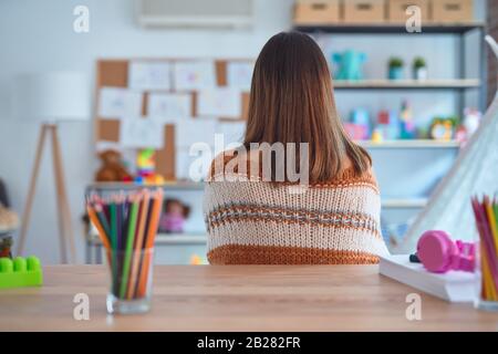 Junge, schöne Lehrerin, die Pullover und Brille trägt, die auf dem Schreibtisch im Kindergarten sitzen und rückwärts mit gekreuzten Armen wegschauen Stockfoto