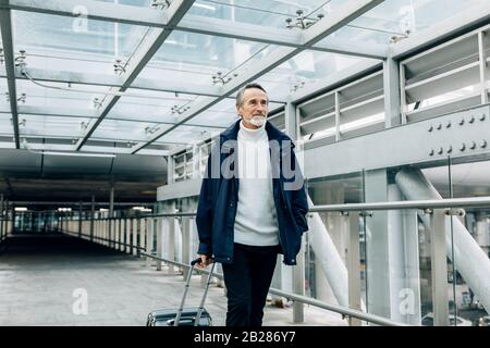 Gutaussehender reifer Mann, der mit seinem Gepäck am Flughafen spazieren ging Stockfoto