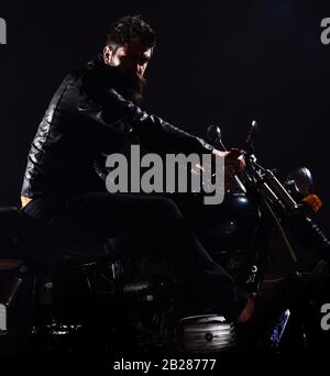 Biker Freizeitkonzept. Macho, brutaler Biker in Lederjacke Motorrad fahren nachts, Platz kopieren. Mann mit Bart, Biker in Lederjacke auf dem Motorrad in Dunkelheit, schwarzer Hintergrund Stockfoto